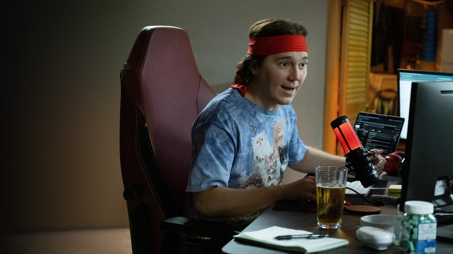 "Dumb Money - Schnelles Geld": Halbnahe: Keith Gill (Paul Dano) sitzt in einem T-Shirt mit Katzenaufdruck und rotem Stirnband an einem vollen Schreibtisch neben einem Glas Bier und spricht mit emotionalem Gesichtsausdruck in ein rot leuchtendes Podcast-Mikrofon. 