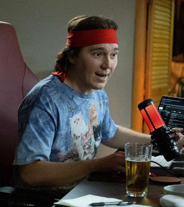 "Dumb Money - Schnelles Geld": Halbnahe: Keith Gill (Paul Dano) sitzt in einem T-Shirt mit Katzenaufdruck und rotem Stirnband an einem vollen Schreibtisch neben einem Glas Bier und spricht mit emotionalem Gesichtsausdruck in ein rot leuchtendes Podcast-Mikrofon. 