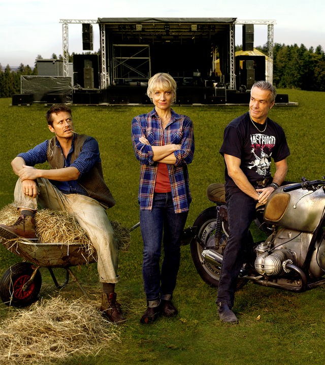  Paul (Roman Knizka), Caro (Christina Große) und Mike (Hannes Jaenicke) stehen auf einer Wiese. Weit im Hintergrund ist die große Bühne zu sehen.