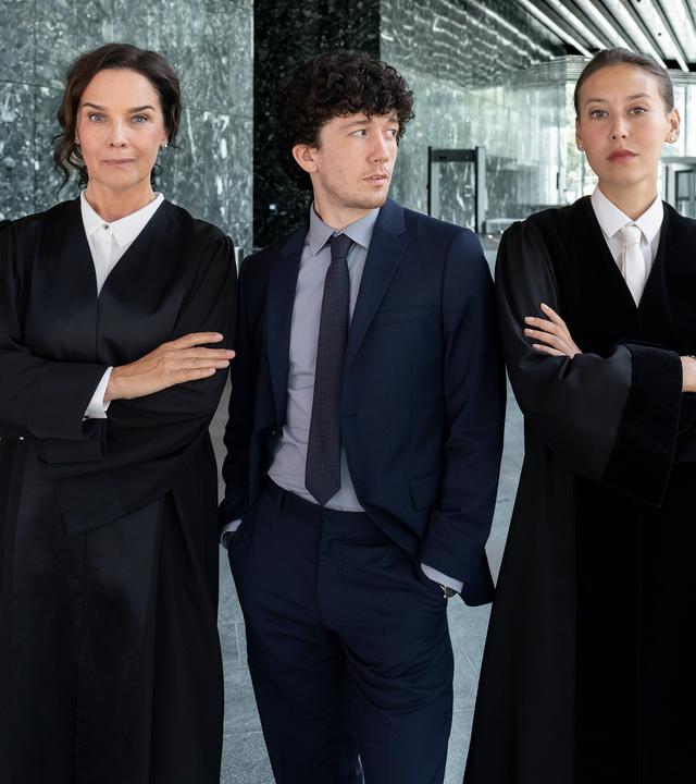 Anna Conti (Désirée Nosbusch), Carlo von Hehenkamp (Maximilian Mundt) und Henry Mahn (Malaya Stern Takeda) stehen nebeneinander im großzügigen Foyer eines modernen Gebäudes.