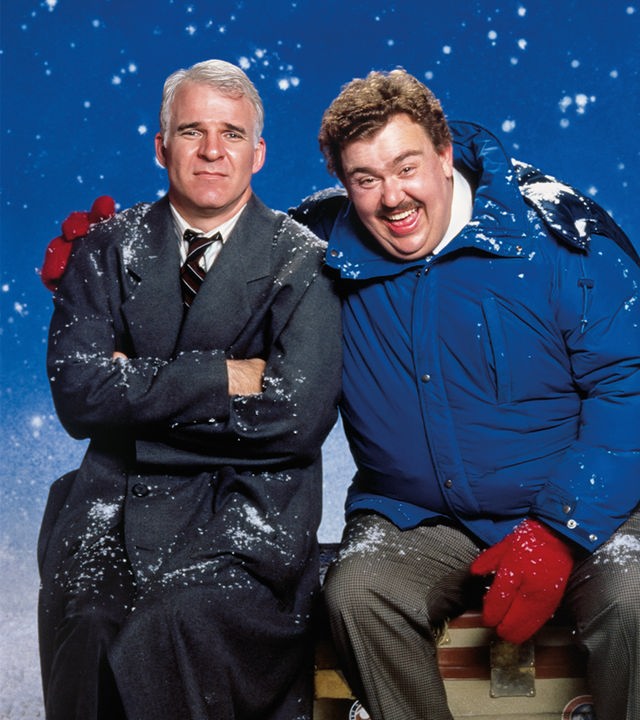 "Ein Ticket für zwei": Neal (Steve Martin) und Del (John Candy) sitzen nebeneinander. Del hat den rechten Arm um Neals Schulter gelegt und lacht. Neal schaut gequält. Im Hintergrund rieseln Schneeflocken.