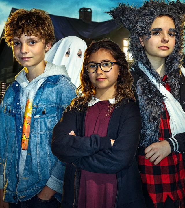 Vier Kinder, eines als Werwolf und eines als Gespenst verkleidet, sind im Vordergrund zu sehen. Es ist dunkel. Im Hintergrund steht ein gruselig beleuchtetes Haus.