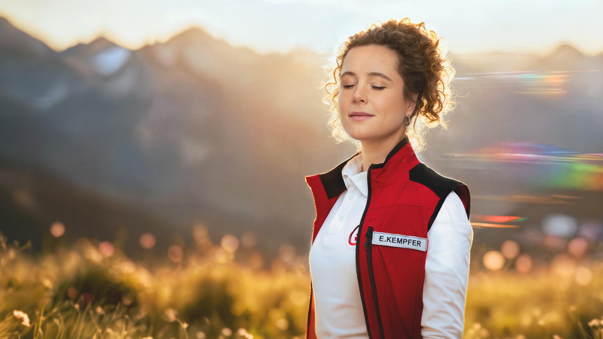 Rettungssanitäterin Elli steht vor der sommerlichen Bergkulisse des Karwendelgebirges. Sie sieht lächelnd den Zuschauer an. Links und rechts von ihrem Kopf sieht man ihr Gesicht noch einmal mit verschiedenen Gesichtsausdrücken - einmal mit geschlossenen Augen und einmal sorgenvoll mit der Hand in ihren Haaren. 