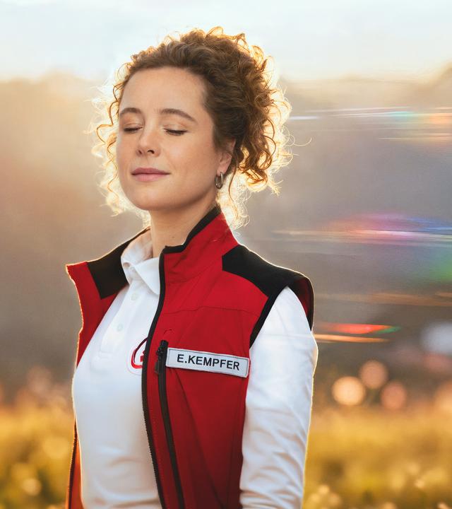 Rettungssanitäterin Elli steht vor der sommerlichen Bergkulisse des Karwendelgebirges. Sie sieht lächelnd den Zuschauer an. Links und rechts von ihrem Kopf sieht man ihr Gesicht noch einmal mit verschiedenen Gesichtsausdrücken - einmal mit geschlossenen Augen und einmal sorgenvoll mit der Hand in ihren Haaren. 