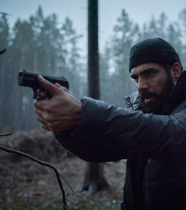 Der Ermittler Assad (Zaki Youssef) befindet sich in einem Waldstück und richtet seine Pistole gegen Angreifer, die auf dem Bild aber nicht zu sehen sind.