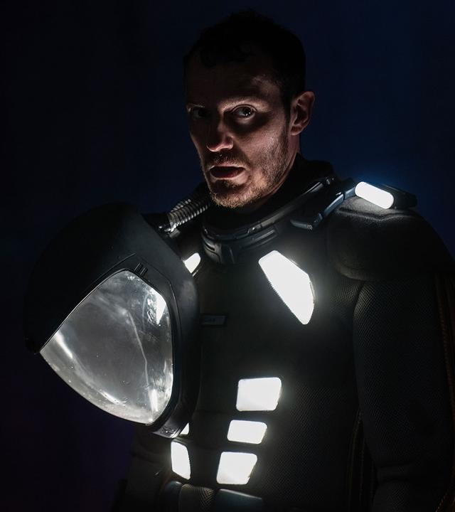 "Eternal": Halbnahe - Elias (Simon Sears) steht in einem Schutzanzug mit weiß leuchtenden Flächen, sowie einem Schutzhelm in einer Hand in absoluter Dunkelheit. Sein angespanntes Gesicht ist zum Teil durch Schatten verborgen.