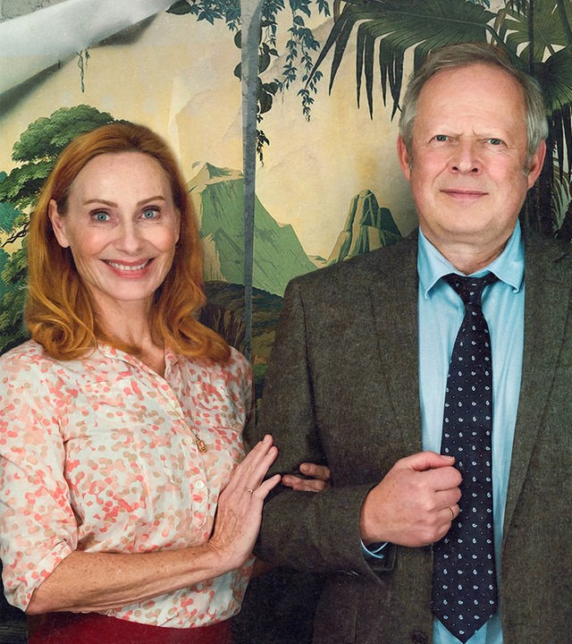 Gundula (Andrea Sawatzki) und Gerald Bundschuh (Axel Milberg) als lächelndes Ehepaar vor einer detailreichen Dschungeltapete. Er trägt ein braunes Sakko mit gemusterter Krawatte, während sie im hellen Oberteil leicht seinen Arm hält.