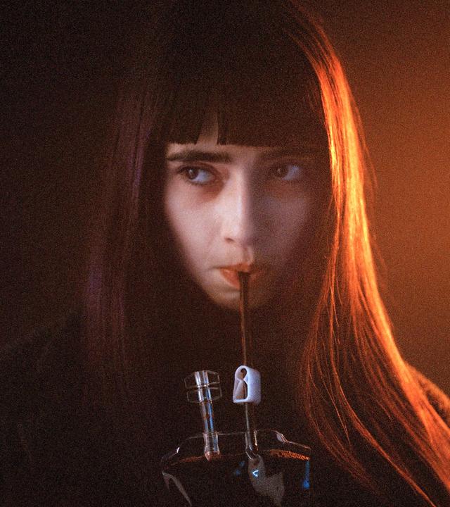 Sasha (Sara Montpetit) trinkt ausdruckslos mit einem Strohhalm aus einem Blutbeutel. Der Hintergrund des Bildes ist schwarz, wobei Sasha von der Seite her von einem grellen, orangefarbenen Licht angestrahlt wird.