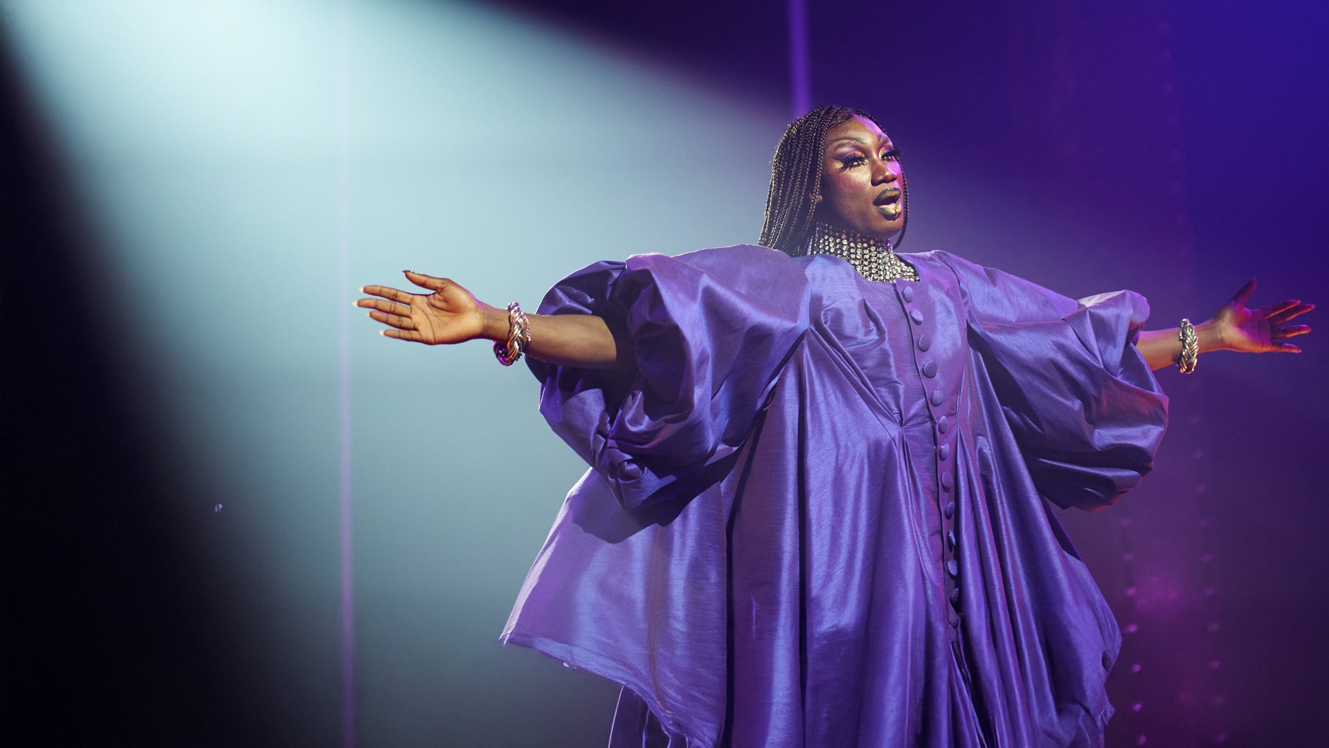 Femme": Halbnahe: Jules (Nathan Stewart-Jarrett) steht als Dragqueen in einem opulentem lila Kleid mit dünn geflochtenen Zöpfen und ausgebreiteten Armen in Scheinwerferlicht auf Bühne. 