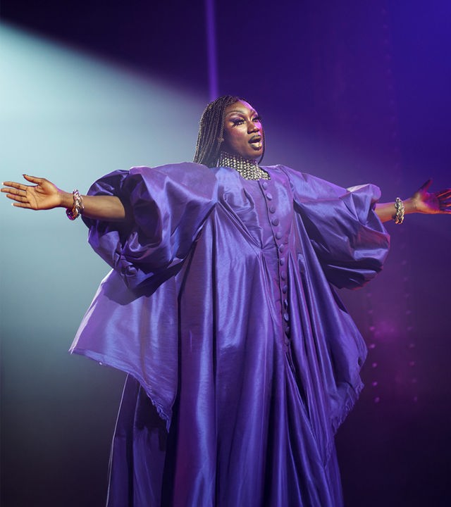 Femme": Halbnahe: Jules (Nathan Stewart-Jarrett) steht als Dragqueen in einem opulentem lila Kleid mit dünn geflochtenen Zöpfen und ausgebreiteten Armen in Scheinwerferlicht auf Bühne. 