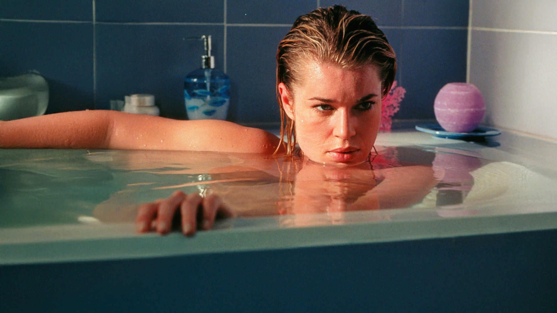 Laure (Rebecca Romijn) liegt in einer hochvollen Badewanne und schaut geheimnisvoll nach halbrechts aus dem Bild. Ihr Kinn ist nur knapp über dem Wasser.