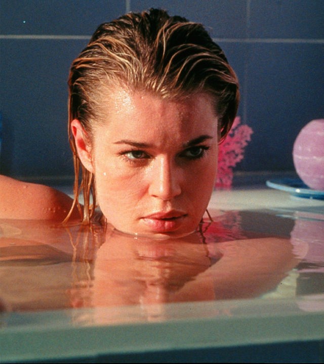 Laure (Rebecca Romijn) liegt in einer hochvollen Badewanne und schaut geheimnisvoll nach halbrechts aus dem Bild. Ihr Kinn ist nur knapp über dem Wasser.