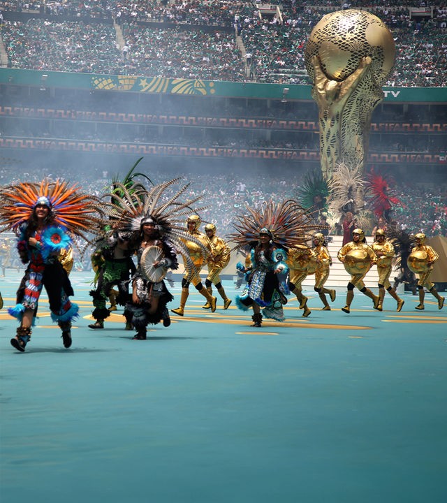 Die Eröffnungsfeier der FIFA Fußball Weltmeisterschaft 2026. Im Stadion in Mexiko City steht ein riesiger WM Pokal um den Menschen in Gewändern der mexikanischen Ureinwohner tanzen und rennen.