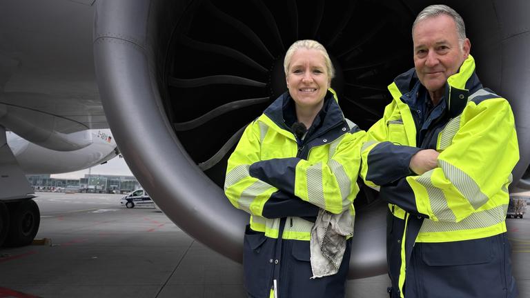 Ein Flugzeugmechaniker und eine Flugzeugmechanikerin stehen in knallgelben Jacken vor einer Turbine eines Jumbojets. Ihre Arme sind verschränkt und sie blicken selbstbewusst in die Kamera.