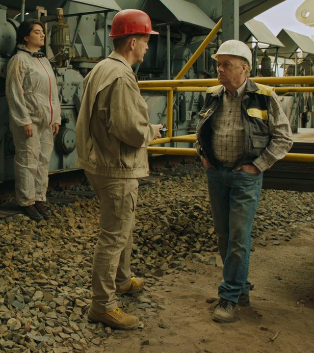 Horàk (Godehard Giese) und Michael (Markus Hering) tragen Schutzhelme und stehen sich vor einem Bagger gegenüber.