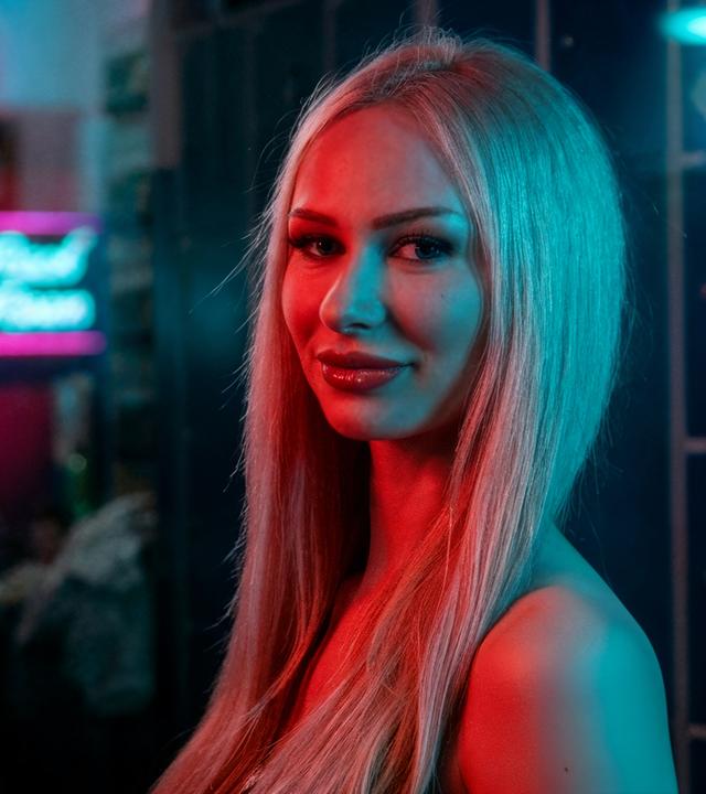 Stripperin Anabelle, 19, tanzt an der Stange des Nachtclubs "Pure Platinum". Die junge Frau mit blonden langen Haaren schaut seitlich in die Kamera. Die Lichteffekte im Hintergrund wurden mit KI bearbeitet.  