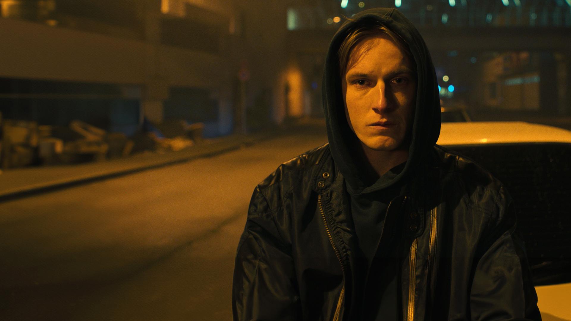 Kai (Louis Hofmann) trägt eine Regenjacke mit Kapuze und steht nachts vor einem Auto auf einer menschenleeren Straße.