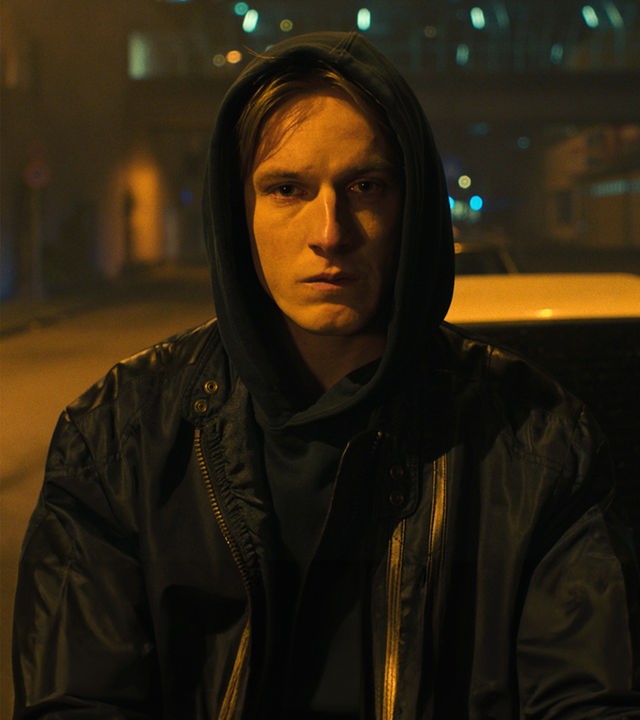 Kai (Louis Hofmann) trägt eine Regenjacke mit Kapuze und steht nachts vor einem Auto auf einer menschenleeren Straße.