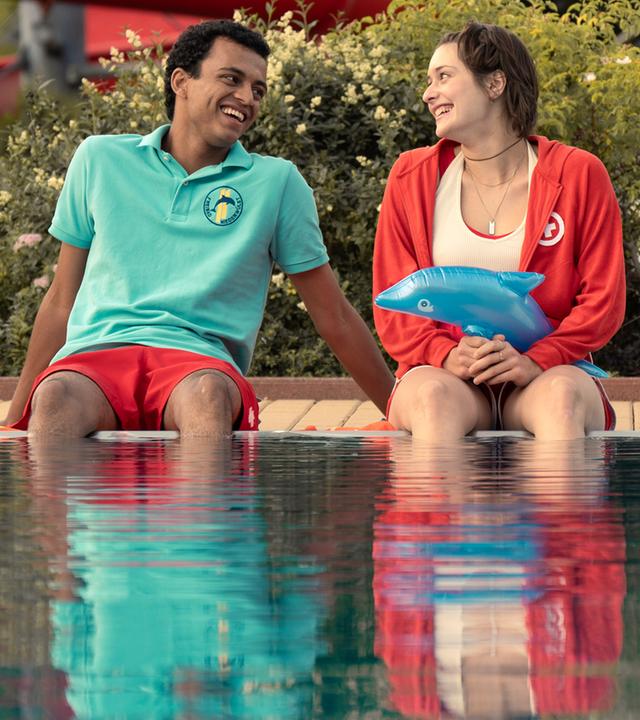 Mira (Anouk Elias) und Simon (Tyrell Otoo) sitzen am dem Becken des Freibads. Beide schauen sich an und lachen. Im Freibad sind keine weiteren Gäste zu sehen.