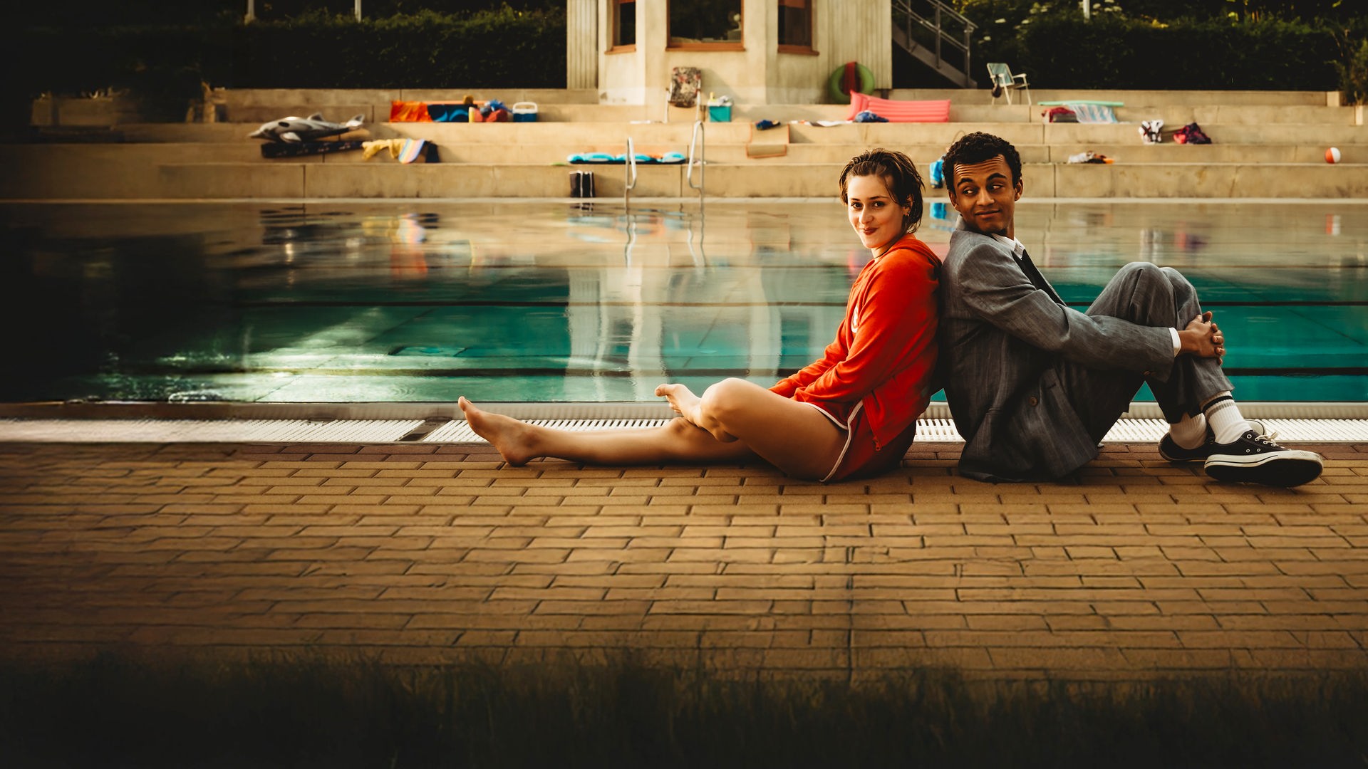 "Für immer Freibad": Mira (Anouk Elias) und Simon (Tyrell Otoo) sitzen Rücken an Rücken vor dem Becken des Freibads. Simon dreht seinen Kopf zu Mira und beide lächeln. Im Freibad sind keine weiteren Gäste zu sehen.