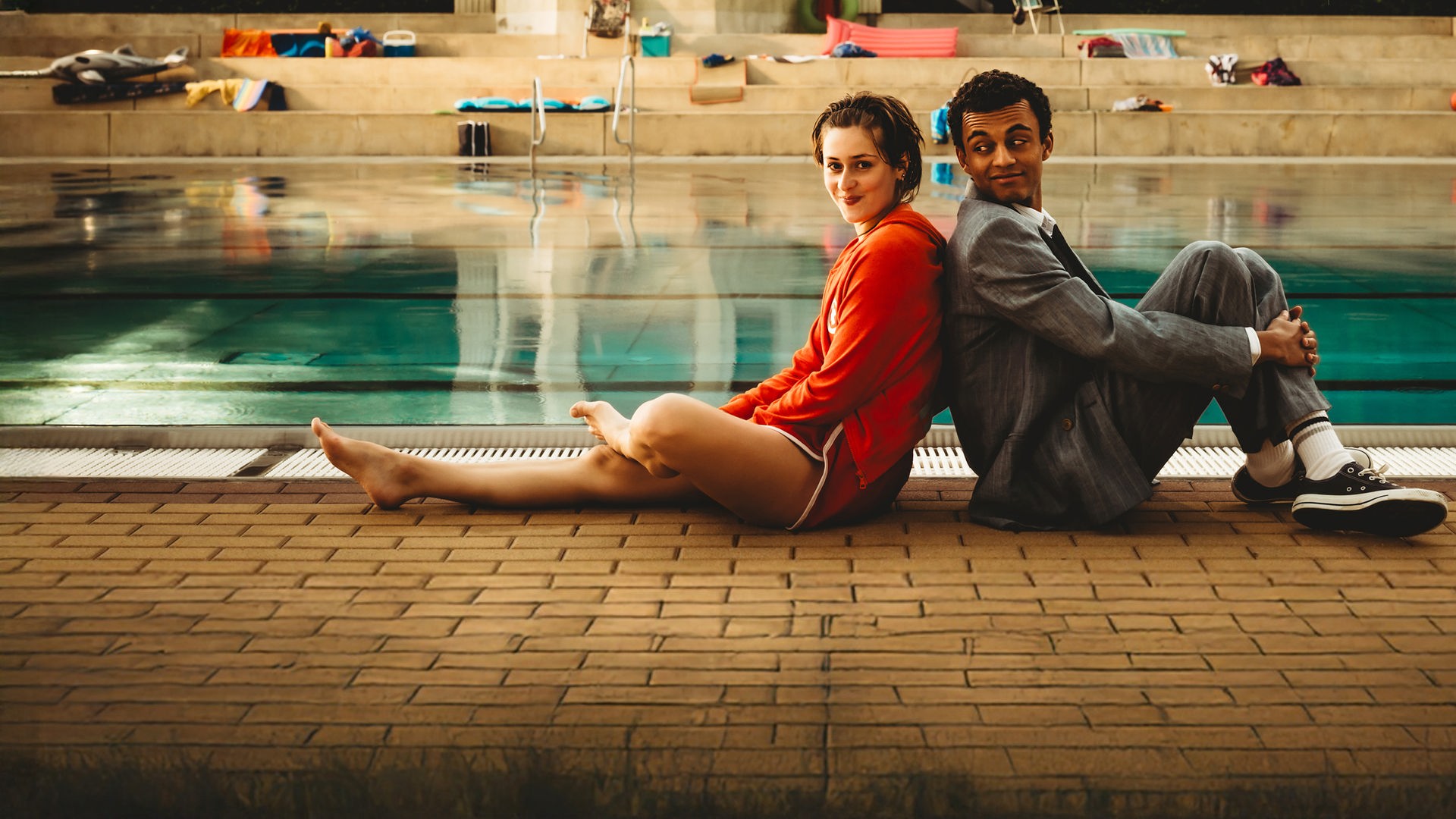 "Für immer Freibad": Mira (Anouk Elias) und Simon (Tyrell Otoo) sitzen Rücken an Rücken vor dem Becken des Freibads. Simon dreht seinen Kopf zu Mira und beide lächeln. Im Freibad sind keine weiteren Gäste zu sehen.