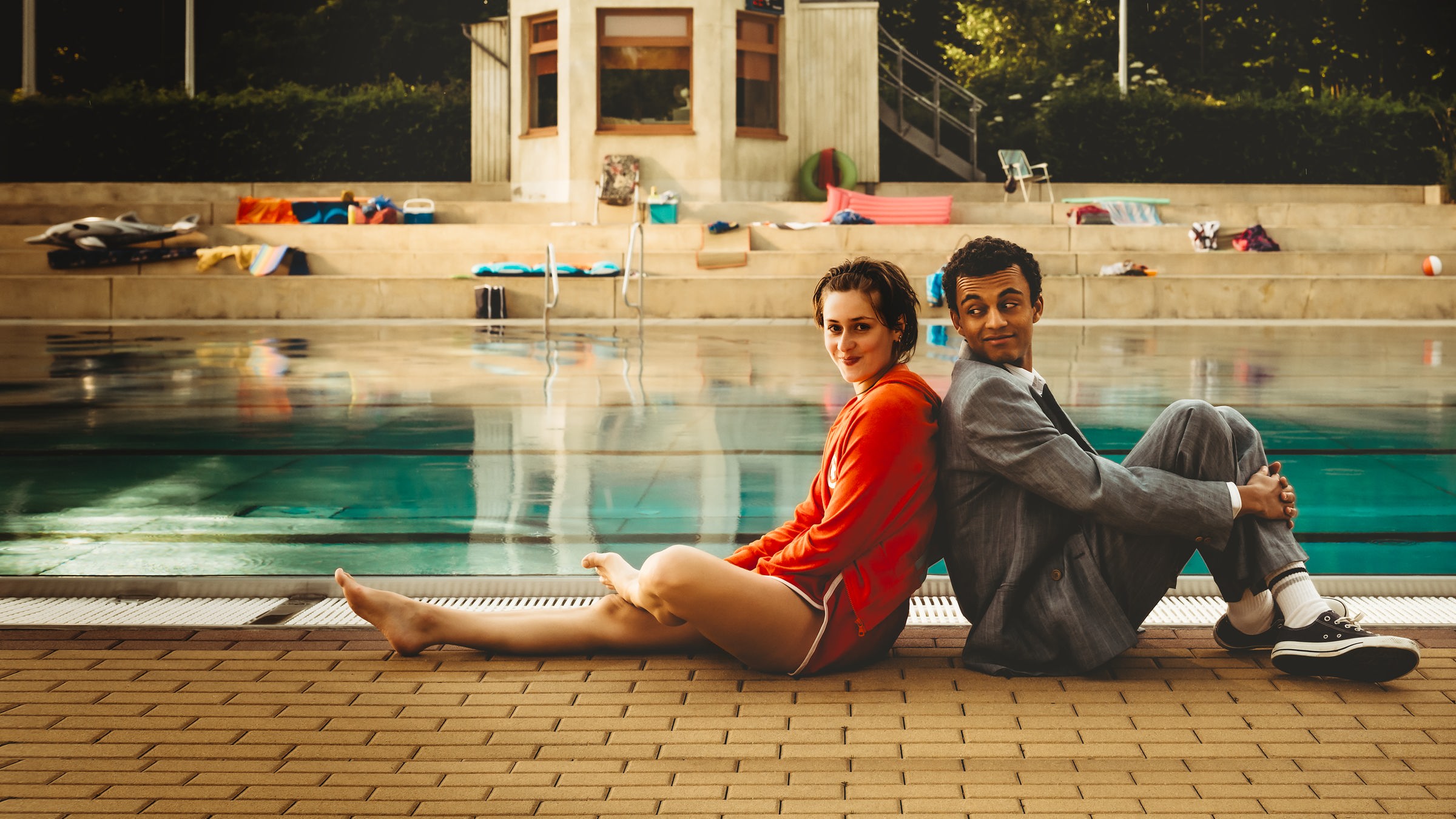 "Für immer Freibad": Mira (Anouk Elias) und Simon (Tyrell Otoo) sitzen Rücken an Rücken vor dem Becken des Freibads. Simon dreht seinen Kopf zu Mira und beide lächeln. Im Freibad sind keine weiteren Gäste zu sehen.