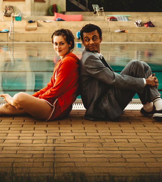 "Für immer Freibad": Mira (Anouk Elias) und Simon (Tyrell Otoo) sitzen Rücken an Rücken vor dem Becken des Freibads. Simon dreht seinen Kopf zu Mira und beide lächeln. Im Freibad sind keine weiteren Gäste zu sehen.