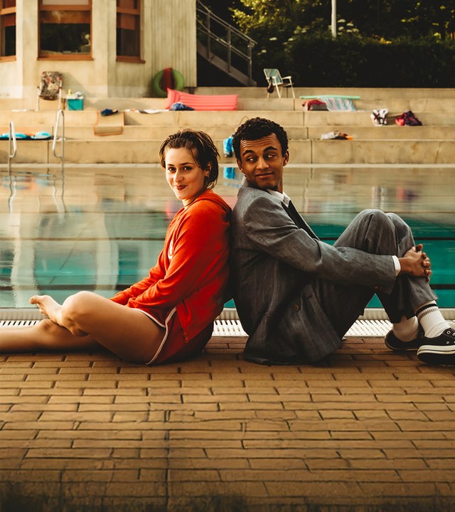 "Für immer Freibad": Mira (Anouk Elias) und Simon (Tyrell Otoo) sitzen Rücken an Rücken vor dem Becken des Freibads. Simon dreht seinen Kopf zu Mira und beide lächeln. Im Freibad sind keine weiteren Gäste zu sehen.