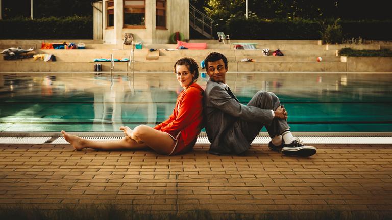 "Für immer Freibad": Mira (Anouk Elias) und Simon (Tyrell Otoo) sitzen Rücken an Rücken vor dem Becken des Freibads. Simon dreht seinen Kopf zu Mira und beide lächeln. Im Freibad sind keine weiteren Gäste zu sehen.