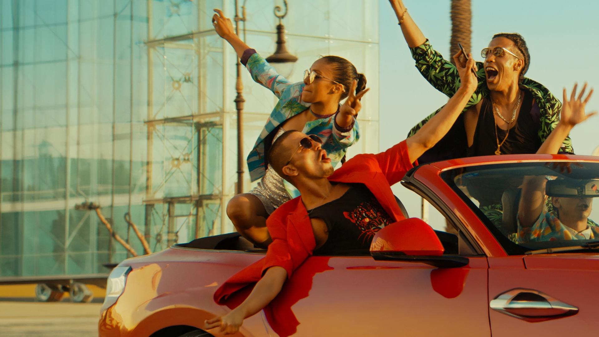 Badia (Nora Gharib), Adamo (Matteo Simoni), Volt (Saïd Boumazoughe) und Junes (Junes Lazaar) fahren in einem roten Cabrio. Alle wirken ausgelassen und strecken feiernd ihre Hände in die Höhe.
