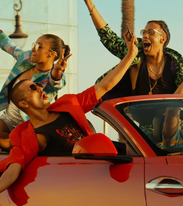 Badia (Nora Gharib), Adamo (Matteo Simoni), Volt (Saïd Boumazoughe) und Junes (Junes Lazaar) fahren in einem roten Cabrio. Alle wirken ausgelassen und strecken feiernd ihre Hände in die Höhe.