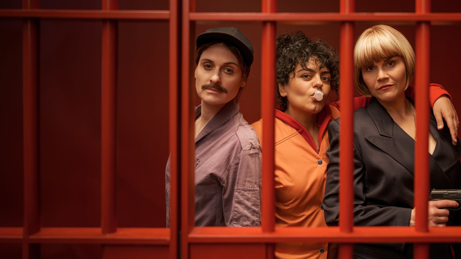 "Gar kein Geld macht auch nicht glücklich": Lesley (Julia Becker), Olivia (Sara Fazilat) und Kim (Katharina Wackernagel) stehen lässig nebeneinander in einem Raum mit knallroten Wänden. Vor ihnen sind rote Gitterstäbe, die die drei Frauen darin einsperren. Olivia formt mit dem Mund eine Kaugummiblase und legt ihren Arm um Kims Schultern. Diese hält eine Pistole in der Hand.
