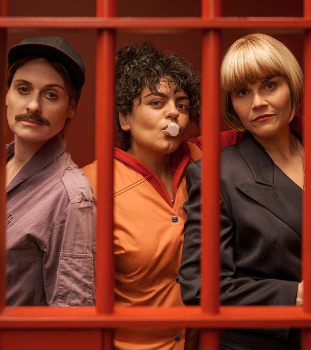 "Gar kein Geld macht auch nicht glücklich": Lesley (Julia Becker), Olivia (Sara Fazilat) und Kim (Katharina Wackernagel) stehen lässig nebeneinander in einem Raum mit knallroten Wänden. Vor ihnen sind rote Gitterstäbe, die die drei Frauen darin einsperren. Olivia formt mit dem Mund eine Kaugummiblase und legt ihren Arm um Kims Schultern. Diese hält eine Pistole in der Hand.