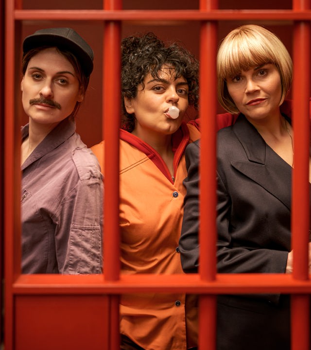 "Gar kein Geld macht auch nicht glücklich": Lesley (Julia Becker), Olivia (Sara Fazilat) und Kim (Katharina Wackernagel) stehen lässig nebeneinander in einem Raum mit knallroten Wänden. Vor ihnen sind rote Gitterstäbe, die die drei Frauen darin einsperren. Olivia formt mit dem Mund eine Kaugummiblase und legt ihren Arm um Kims Schultern. Diese hält eine Pistole in der Hand.