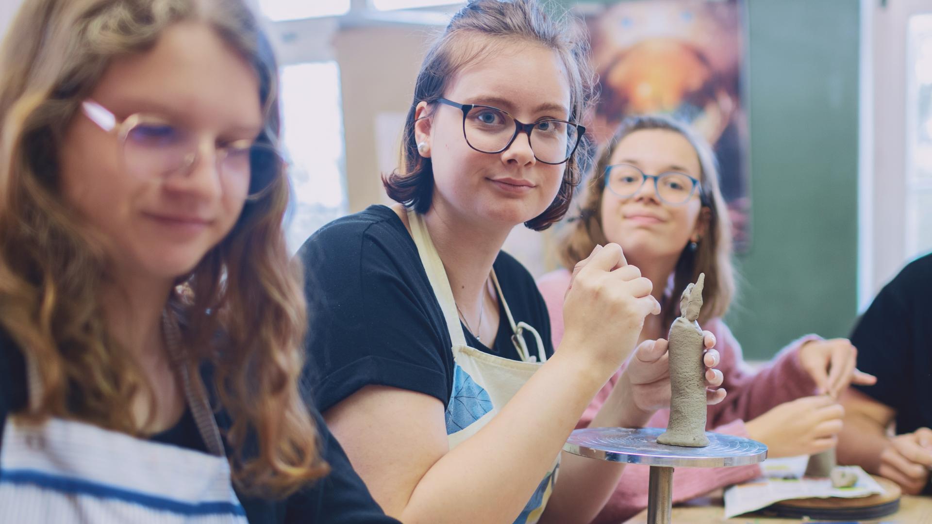 Drei junge Frauen und ein junger Mann sitzen in einem Raum am Tisch. Vor ihnen stehen Töpferscheiben, sie hantieren mit Ton.