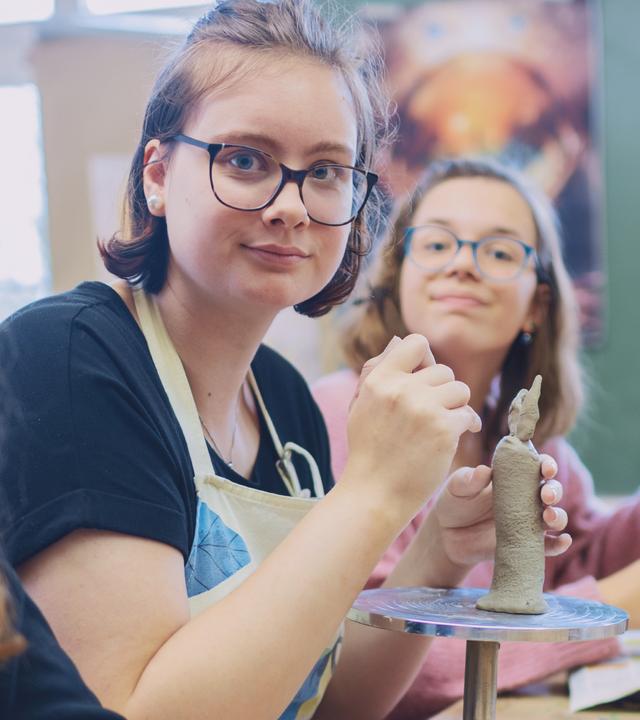 Drei junge Frauen und ein junger Mann sitzen in einem Raum am Tisch. Vor ihnen stehen Töpferscheiben, sie hantieren mit Ton.