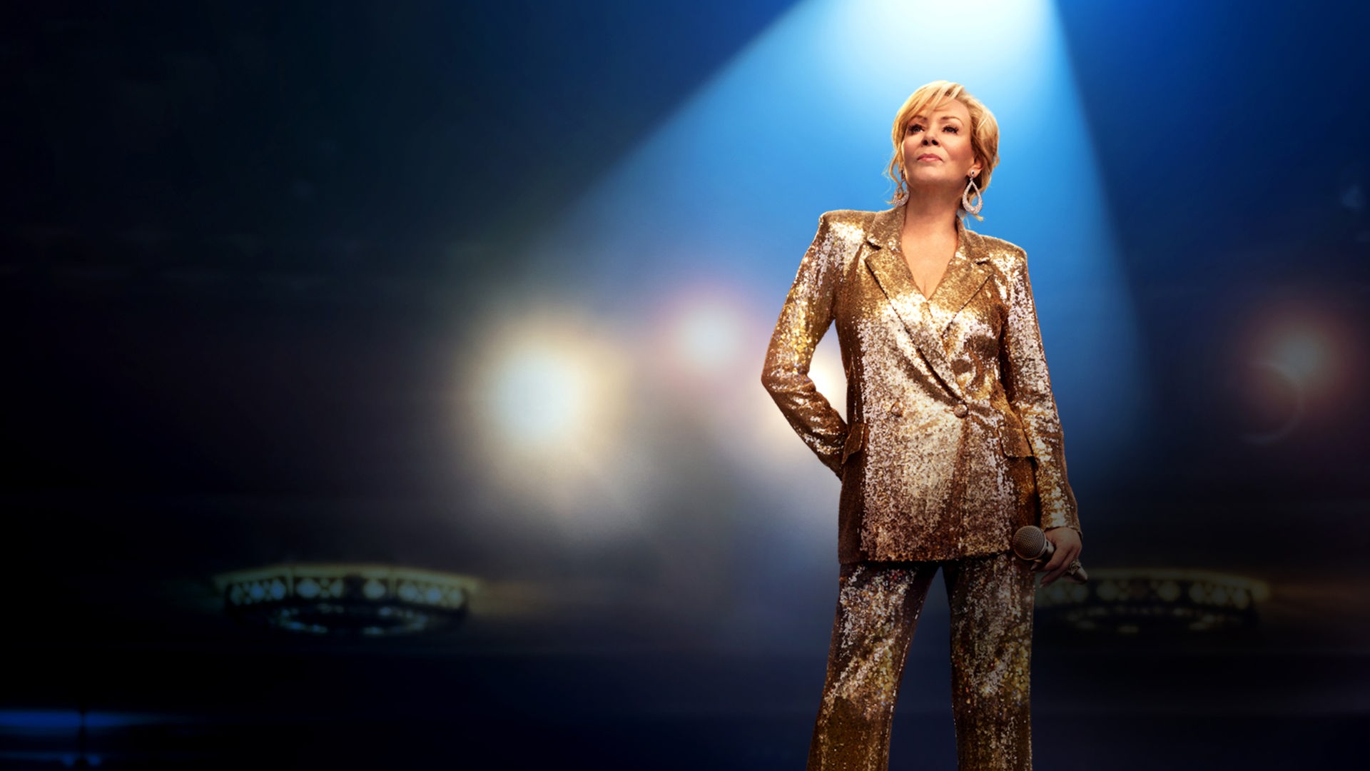 Hacks Staffel 1: Die alternde Komikerin Deborah Vance (Jean Smart) steht in einem glitzernden, goldenen Hosenanzug selbstbewusst im Rampenlicht auf einer Bühne vor dunklem Hintergrund.