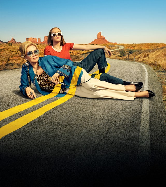 Hacks Staffel 2: Deborah Vance (Jean Smart) und ihre junge Autorin Ava Daniels (Hannah Einbinder) posieren lässig vor einer weiten Wüstenstraße. Sie tragen Sonnenbrillen und lehnen sich aneinander, was eine Aufbruchsstimmung für ihren gemeinsamen Roadtrip vermittelt.