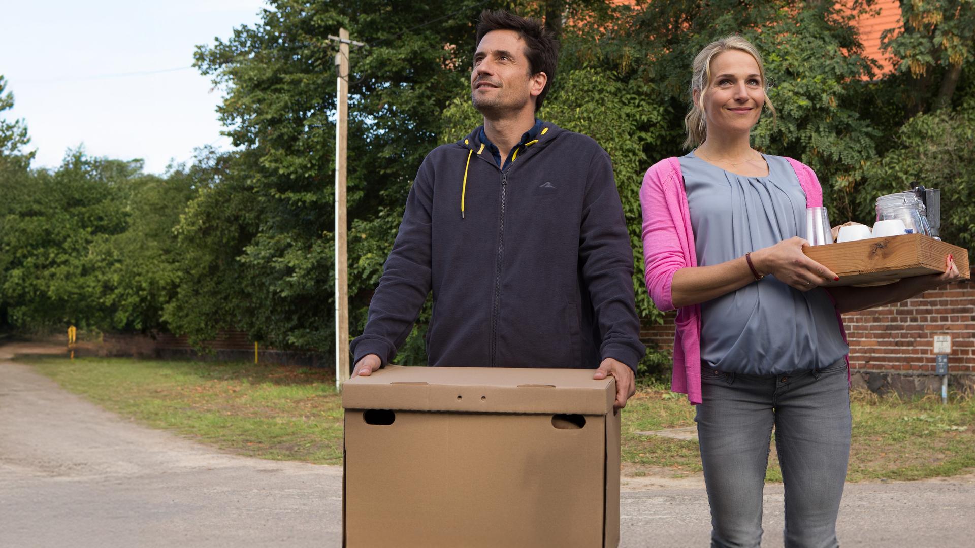 Stefan (Oliver Mommsen, li.) trägt einen Umzugskarton und die schwangere Silke (Tanja Wedhorn, re.) ein Tablett mit Geschirr.