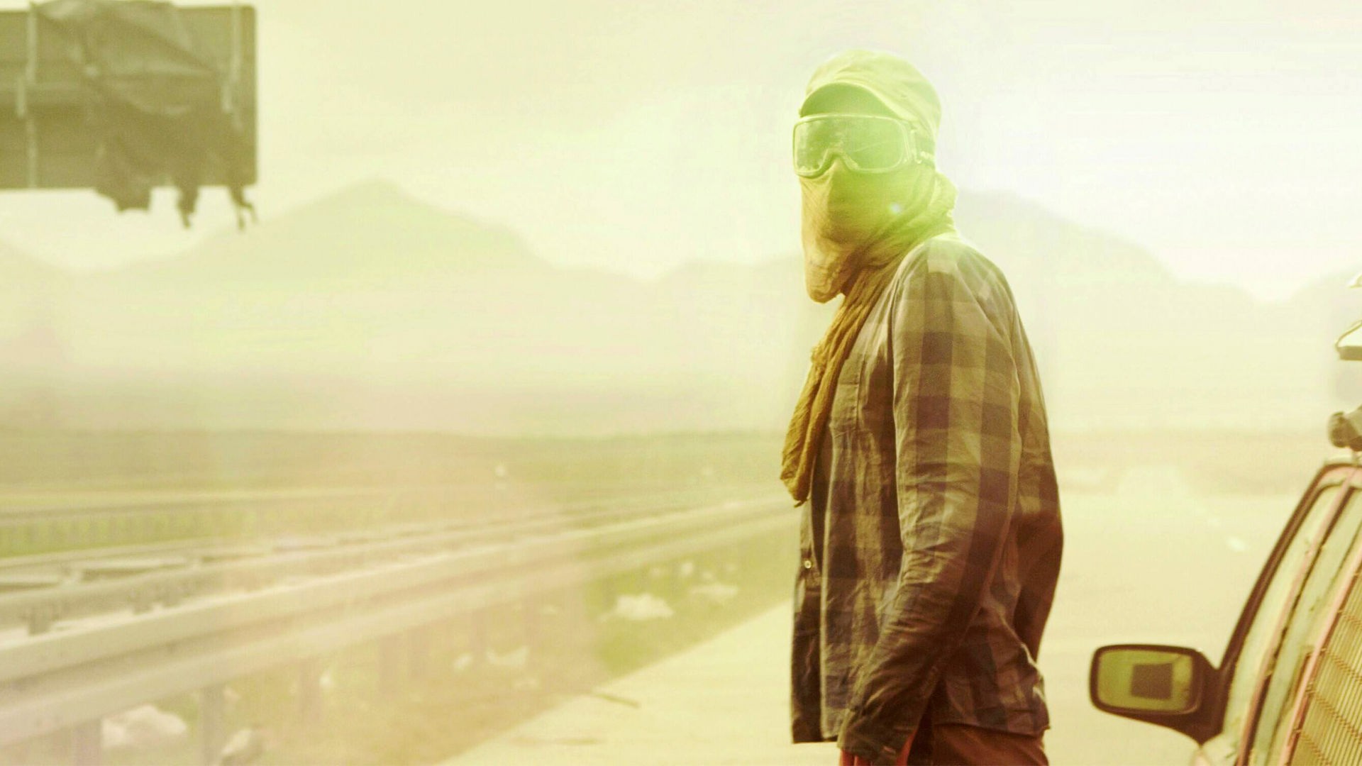 "Hell": Halbnahe Gegenlichtaufnahme mit leichtem Gelbfilter: Eine vermummte Person, mit Schildkappe, Skisonnenbrille und Tuch vor Mund, steht neben einem Auto auf einer verlassenen und zugemüllten Schnellstraße.