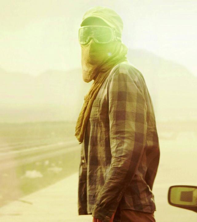 "Hell": Halbnahe Gegenlichtaufnahme mit leichtem Gelbfilter: Eine vermummte Person, mit Schildkappe, Skisonnenbrille und Tuch vor Mund, steht neben einem Auto auf einer verlassenen und zugemüllten Schnellstraße.