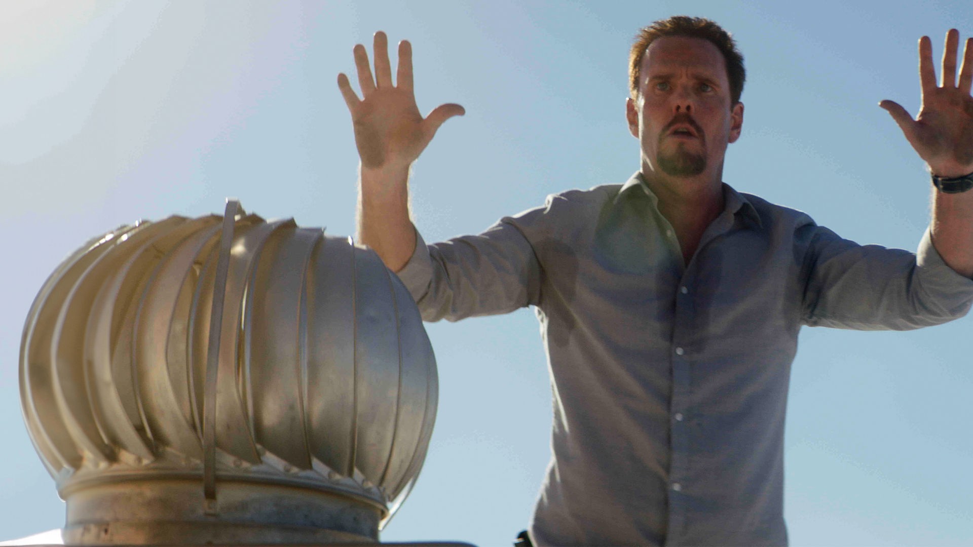 "Hot Seat": Halbnahe, leichte Untersicht: Orlando (Kevin Dillon) steht mit erhobenen Händen und Schweißflecken auf seinem Hemd in der prallen Sonne neben einer runden, metallfarbenen Skulptur und blickt angespannt auf etwas vor ihm.