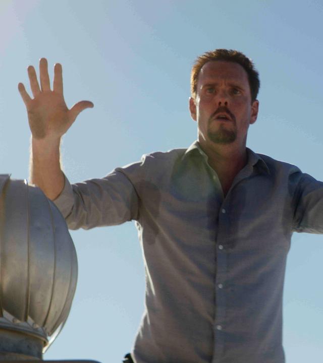 "Hot Seat": Halbnahe, leichte Untersicht: Orlando (Kevin Dillon) steht mit erhobenen Händen und Schweißflecken auf seinem Hemd in der prallen Sonne neben einer runden, metallfarbenen Skulptur und blickt angespannt auf etwas vor ihm.