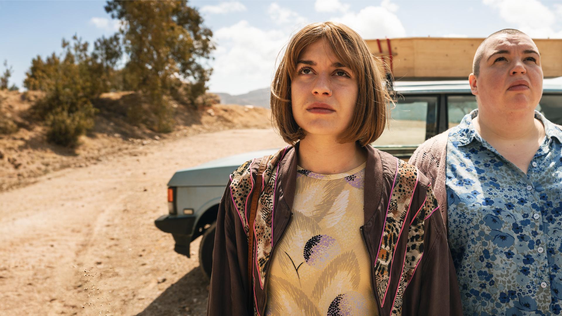 Anna (Emma Rotsaert) und ihre Schwester Kat (Marjan De Schutter) stehen nebeneinander vor ihrem alten Landrover. Sie schauen in den Himmel. Auf dem Dach ihres Autos ist ein einfacher Holzsarg befestigt.