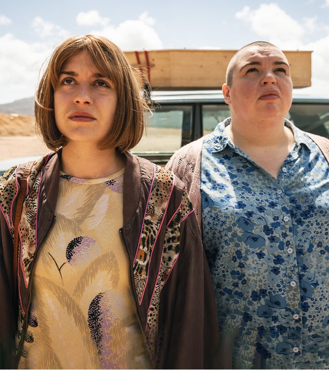 Anna (Emma Rotsaert) und ihre Schwester Kat (Marjan De Schutter) stehen nebeneinander vor ihrem alten Landrover. Sie schauen in den Himmel. Auf dem Dach ihres Autos ist ein einfacher Holzsarg befestigt.