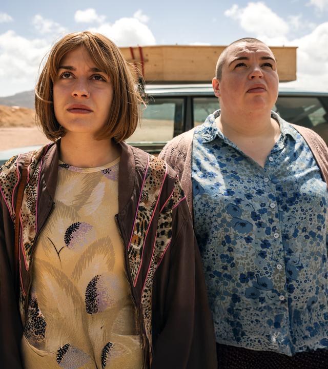 Anna (Emma Rotsaert) und ihre Schwester Kat (Marjan De Schutter) stehen nebeneinander vor ihrem alten Landrover. Sie schauen in den Himmel. Auf dem Dach ihres Autos ist ein einfacher Holzsarg befestigt.