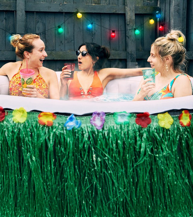 "Hullraisers - Breadcakes": Toni (Leah Brotherhead), Rana (Taj Atwal) und Paula (Sinead Matthews) sitzen zusammen im Bikini in einem aufblasbaren Swimmingpool und schauen sich lachend an, während sie etwas trinken.
