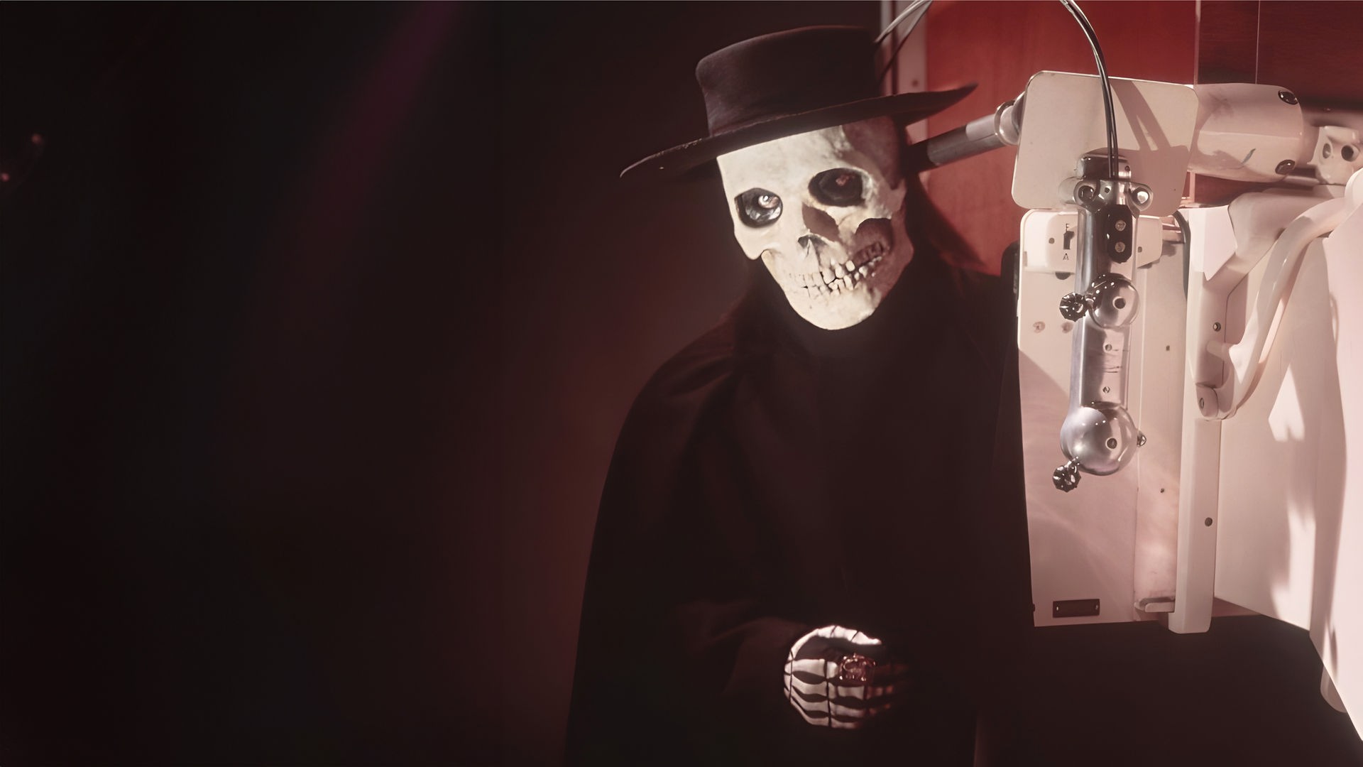 Ein Mann in einer Totenkopfmaske mit einem schwarzen Hut steht hinter einem medizinischen Gerät, das ein Röntgenbild seines Brustkorbs zeigt, auf dem ein Skelett deutlich zu erkennen ist.
