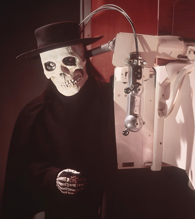 Ein Mann in einer Totenkopfmaske mit einem schwarzen Hut steht hinter einem medizinischen Gerät, das ein Röntgenbild seines Brustkorbs zeigt, auf dem ein Skelett deutlich zu erkennen ist.