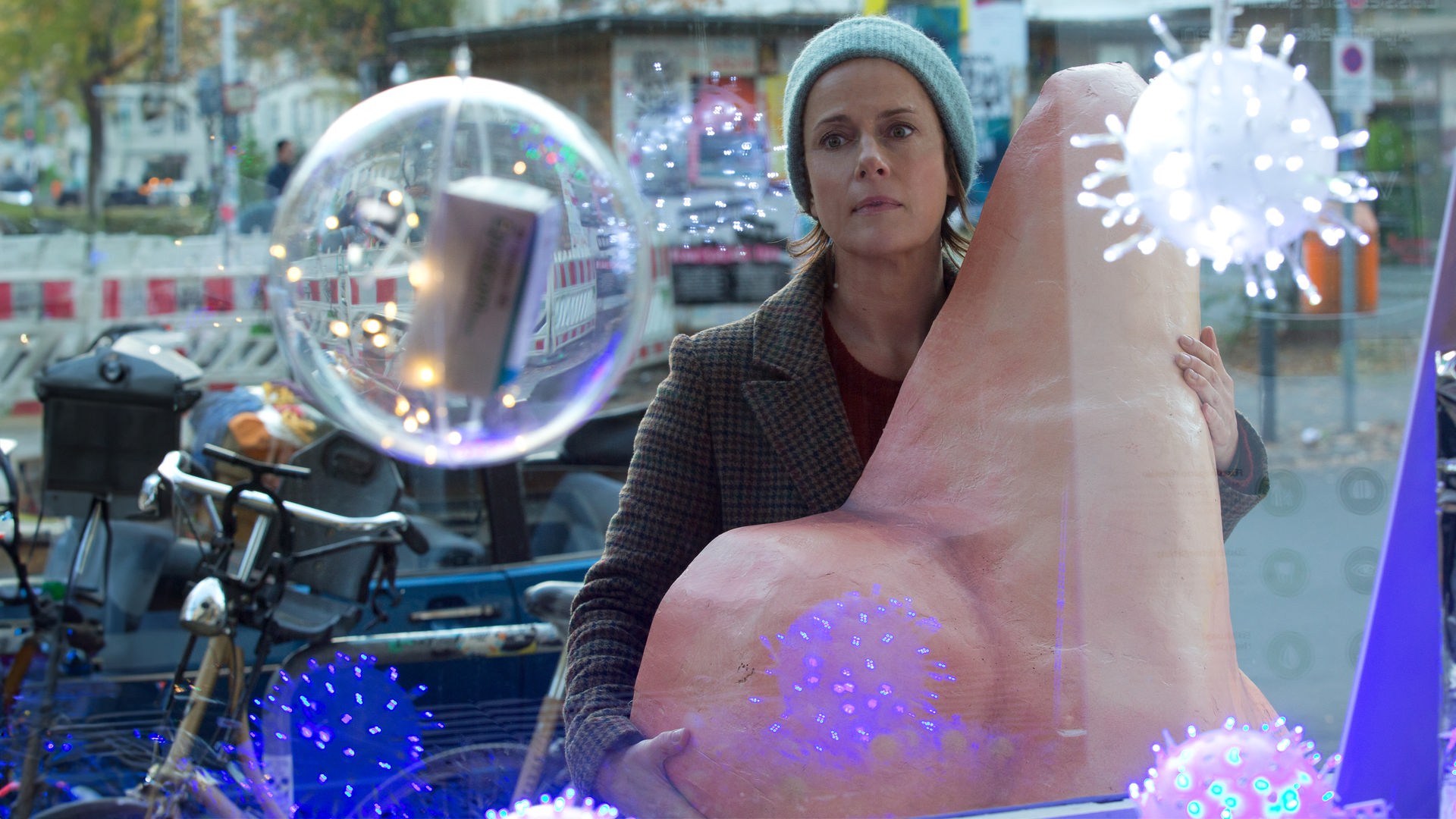 Vor dem Schaufenster einer Apotheke steht Tanja (Claudia Michelsen) mit einer Pappmaché-Nase. Im Fenster blinken bunte Lichtkugeln, die wie große Viren aussehen.